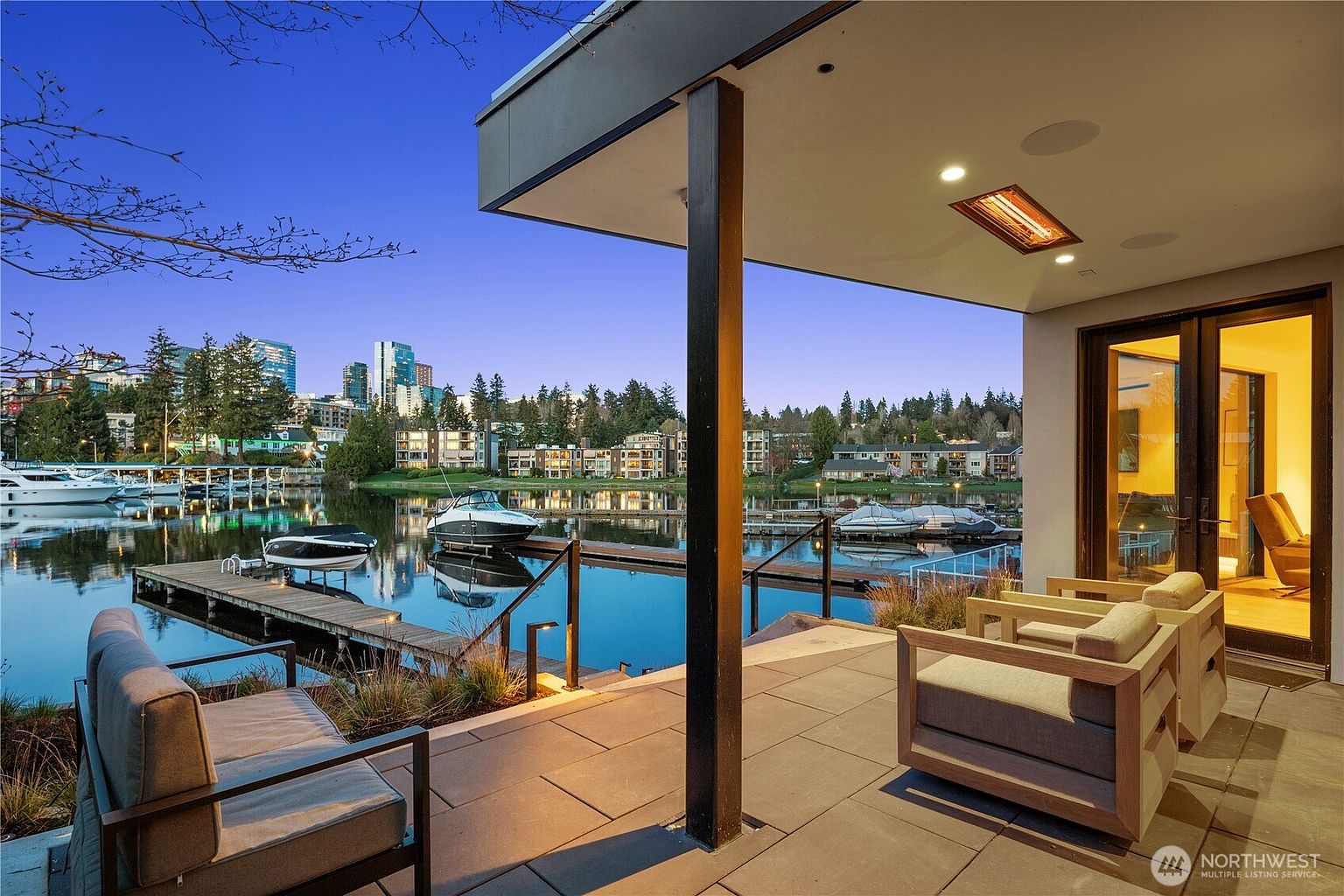 This covered patio offers a serene, elevated view of a calm waterfront with private boat slips and a distant city skyline at dusk. The space is furnished with comfortable, modern outdoor seating and features a built-in ceiling heater for year-round enjoyment. Large glass doors provide a seamless transition between the indoor living area and this tranquil outdoor retreat.