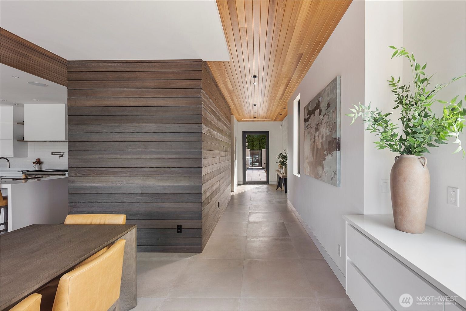 This modern hallway features a striking contrast between a dark, horizontal wood-paneled accent wall and smooth, light-colored walls. A warm wood-slat ceiling adds architectural interest, leading the eye toward a glass-paned door at the end of the corridor that opens to an outdoor space. To the right, a sleek white console table holds a large decorative vase with greenery, while a dining area with yellow chairs is partially visible on the left.