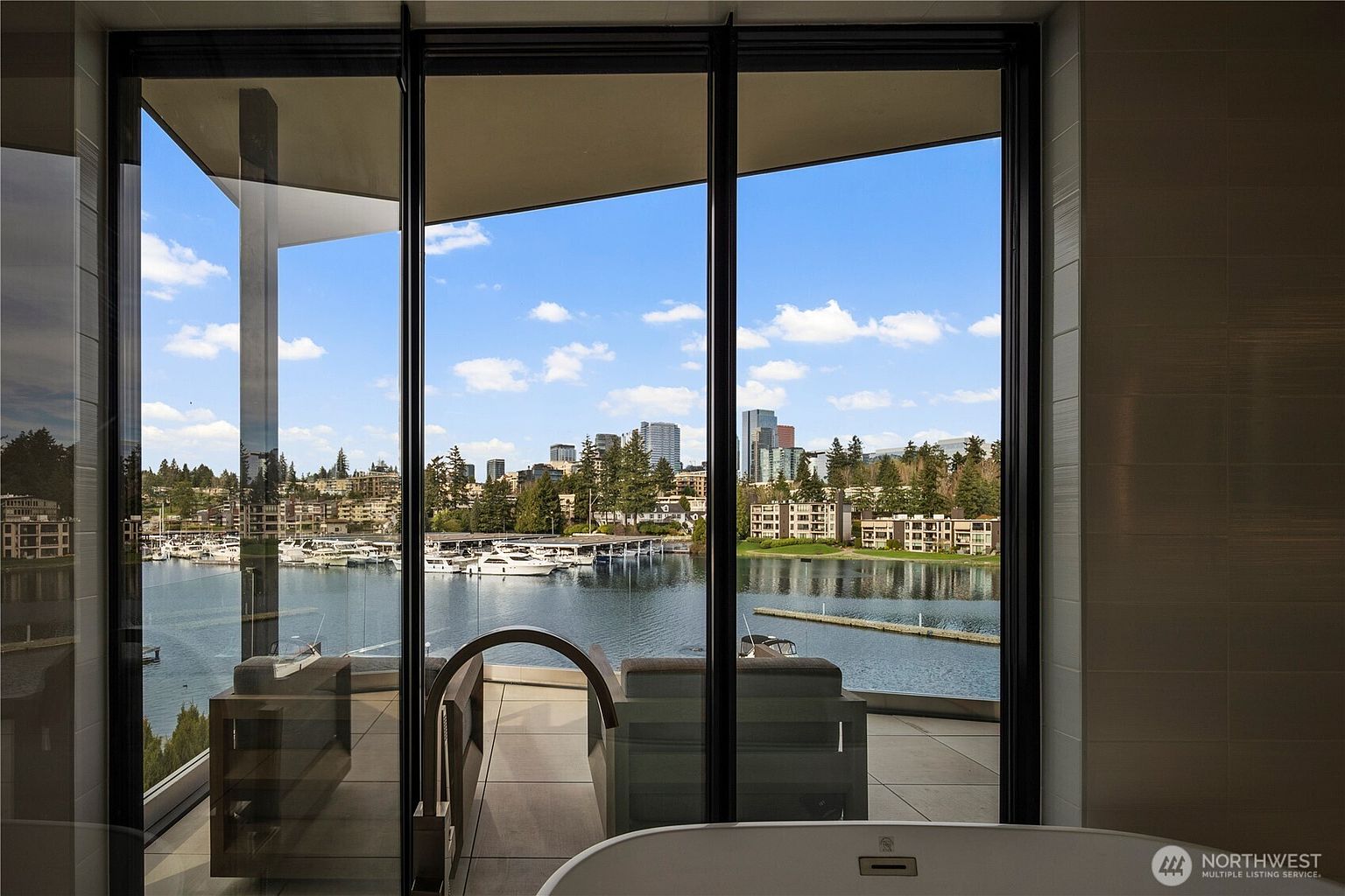 This interior view from a bathroom features a sleek, modern soaking tub positioned in front of expansive floor-to-ceiling windows that offer a panoramic view of a marina and city skyline. The perspective looks out onto a private balcony with outdoor seating, seamlessly blending the indoor space with the waterfront scenery. The clean lines and minimalist aesthetic emphasize the luxurious, tranquil atmosphere of the home.