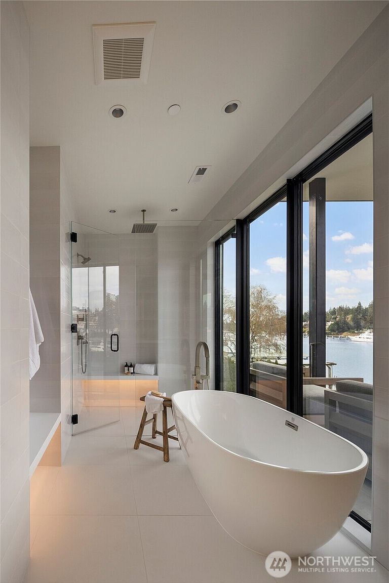 This luxurious primary bathroom features a sleek, freestanding soaking tub positioned next to expansive floor-to-ceiling windows that offer a serene waterfront view. A glass-enclosed walk-in shower with a rainfall showerhead and integrated bench seating sits adjacent to the tub, while a small wooden stool adds a touch of organic warmth to the minimalist, light-toned space. The room is illuminated by recessed lighting and soft under-cabinet glow, creating a tranquil and spa-like atmosphere.
