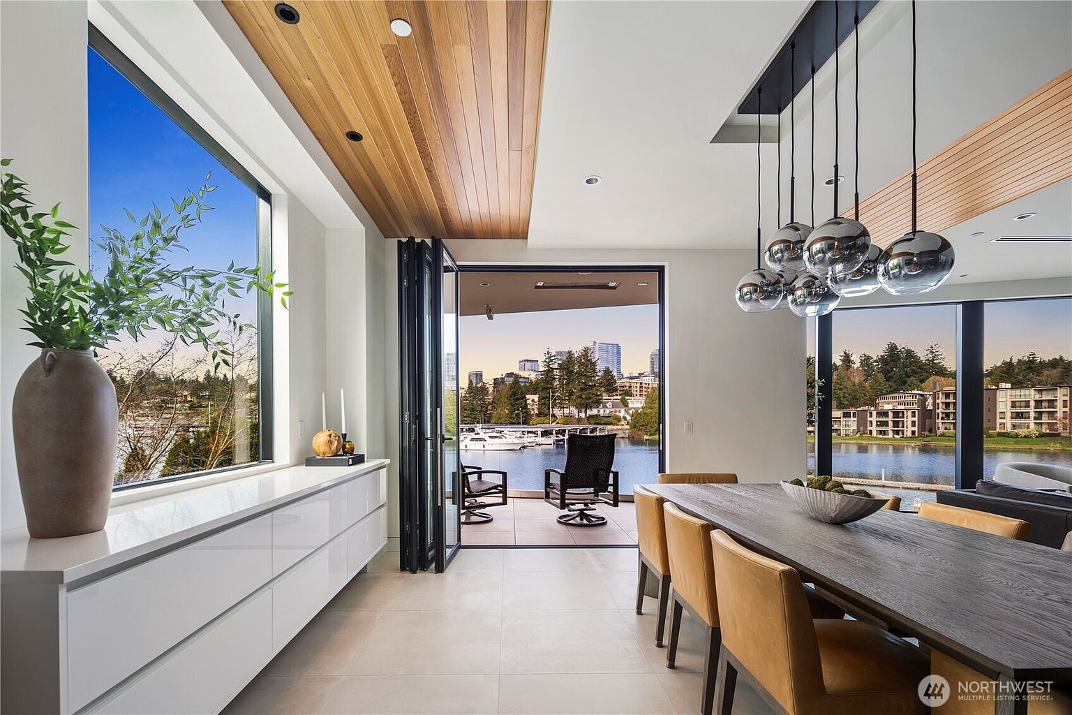 This contemporary dining room features a long, dark wood table surrounded by tan upholstered chairs, illuminated by a cluster of modern spherical pendant lights. The space seamlessly transitions to an outdoor patio through a set of folding glass doors, offering a scenic view of the water and city skyline. A sleek, white built-in console runs along the left wall, topped with a large decorative vase, while a wood-paneled ceiling adds warmth to the bright, open-concept interior.