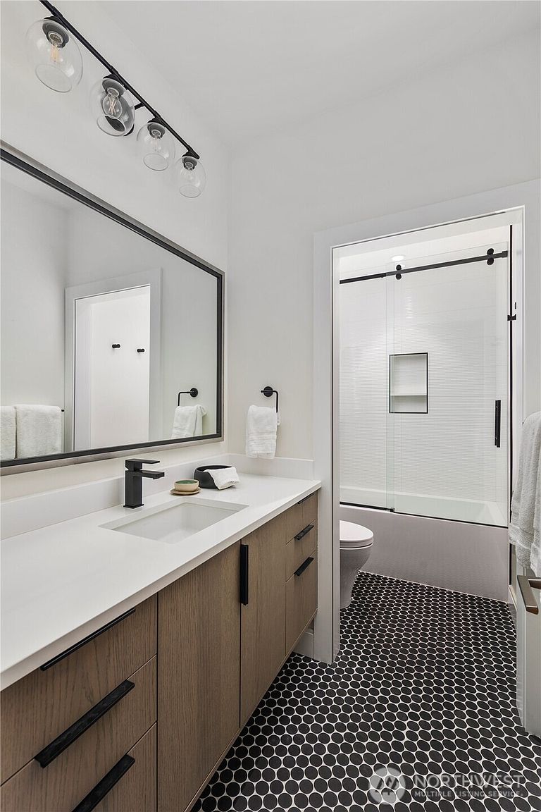This modern bathroom features a vanity with light wood cabinetry, a white countertop, and a black faucet, complemented by a large rectangular mirror and a black light fixture. The floor is covered in a distinctive black and white penny tile pattern, which contrasts with the clean white walls. A glass-enclosed shower and tub combination is visible through the doorway, adding to the contemporary and functional design of the space.