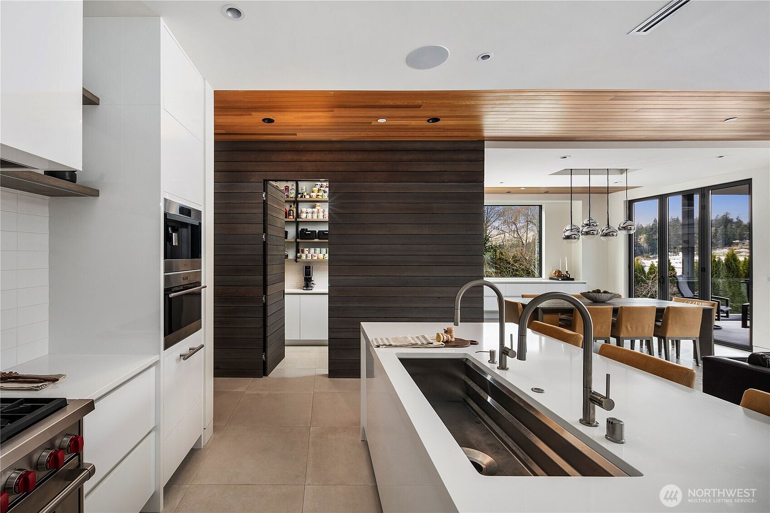 This modern kitchen features a large central island with a deep stainless steel sink and dual faucets, set against a backdrop of sleek white cabinetry and a hidden pantry entrance. The space is defined by a striking contrast between the white surfaces and the dark, horizontal wood-paneled wall that leads into the pantry. Beyond the kitchen, a dining area with warm-toned chairs and a view of the outdoors creates an inviting, open-concept atmosphere.