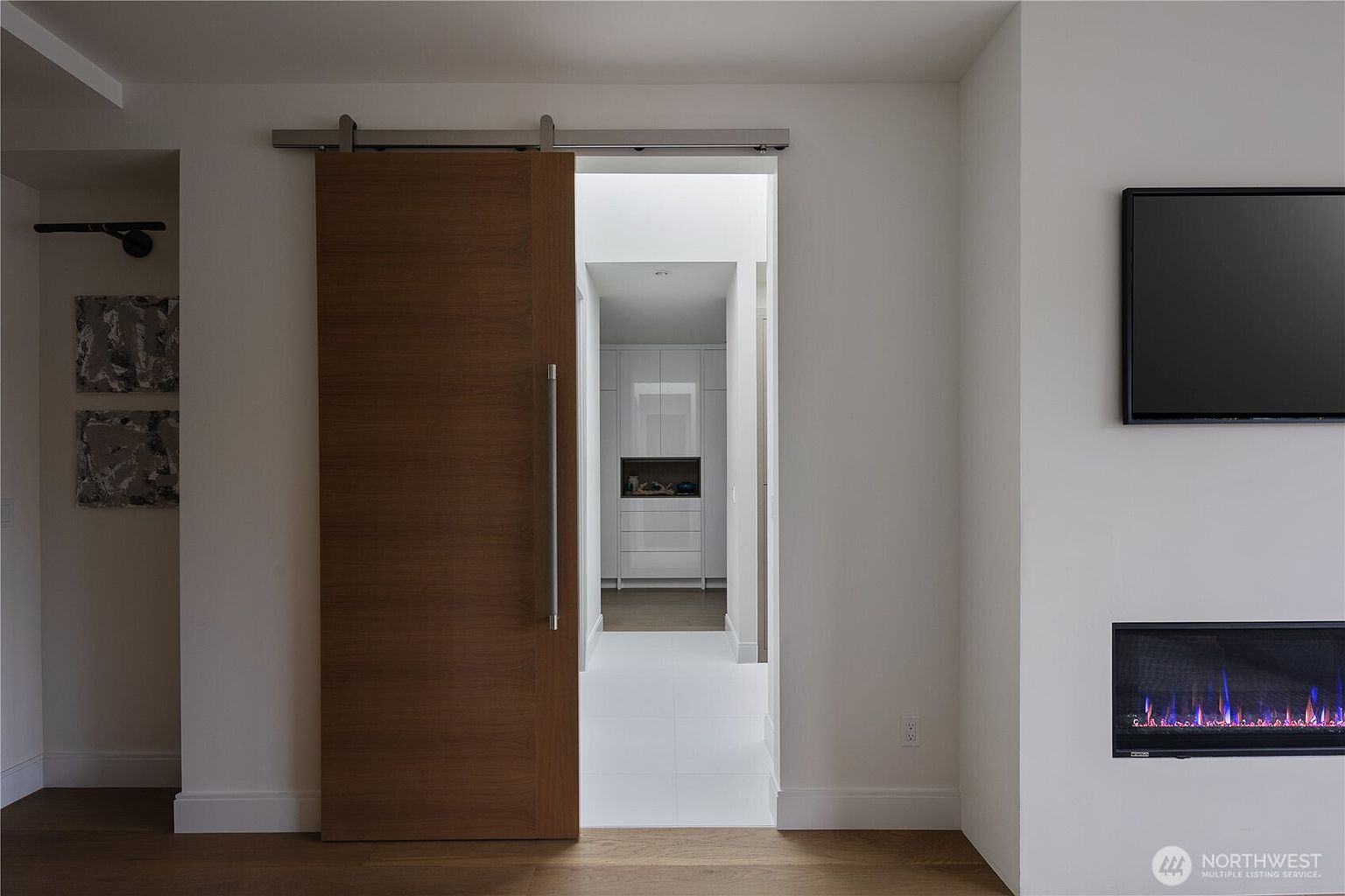 This modern living space features a sleek, wall-mounted electric fireplace with a vibrant blue flame effect and a television mounted above it. A large, wood-toned sliding barn door serves as a prominent architectural feature, opening into a bright hallway with white cabinetry. The room is finished with light-toned wood flooring and crisp white walls, creating a clean and contemporary aesthetic.