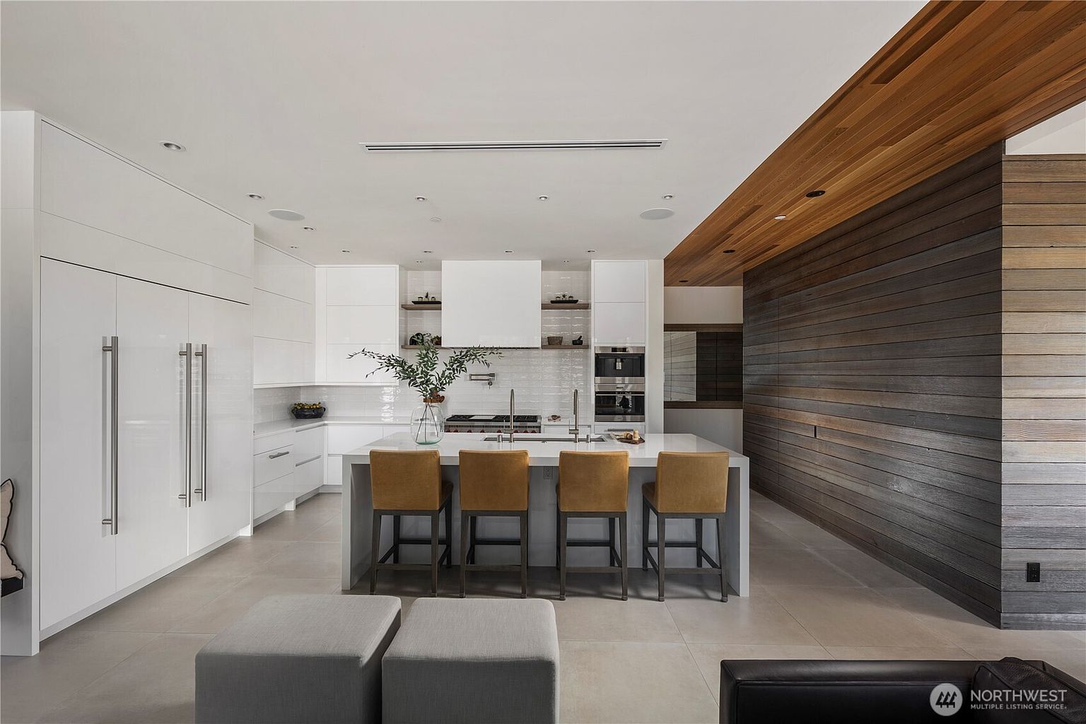 This modern kitchen features a large central island with four upholstered bar stools, set against a backdrop of sleek white cabinetry and a tiled backsplash. A striking wood-paneled wall adds warmth to the space, extending into a ceiling feature that contrasts with the minimalist white design. The perspective is a straight-on, eye-level shot that highlights the open-concept layout and the clean, contemporary aesthetic.