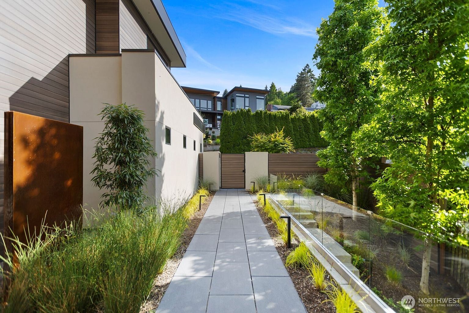 This modern entryway features a clean, paved walkway leading toward a wooden gate, flanked by a minimalist light-colored wall and a glass-railed garden bed. The landscaping includes ornamental grasses and a vibrant green tree, creating a sophisticated and private transition to the home. The perspective is a ground-level shot looking down the path, emphasizing the architectural lines and the contemporary aesthetic of the property.