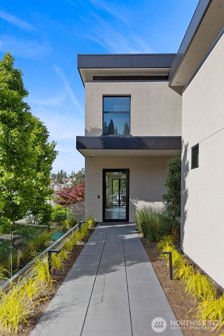 This modern entryway features a clean, minimalist aesthetic with a paved walkway leading to a glass-paneled front door. The structure is characterized by smooth, light-toned walls, dark trim, and a prominent upper-level window that adds architectural depth. Landscaped garden beds with ornamental grasses line the path, while a glass railing provides a clear view of the surrounding landscape.