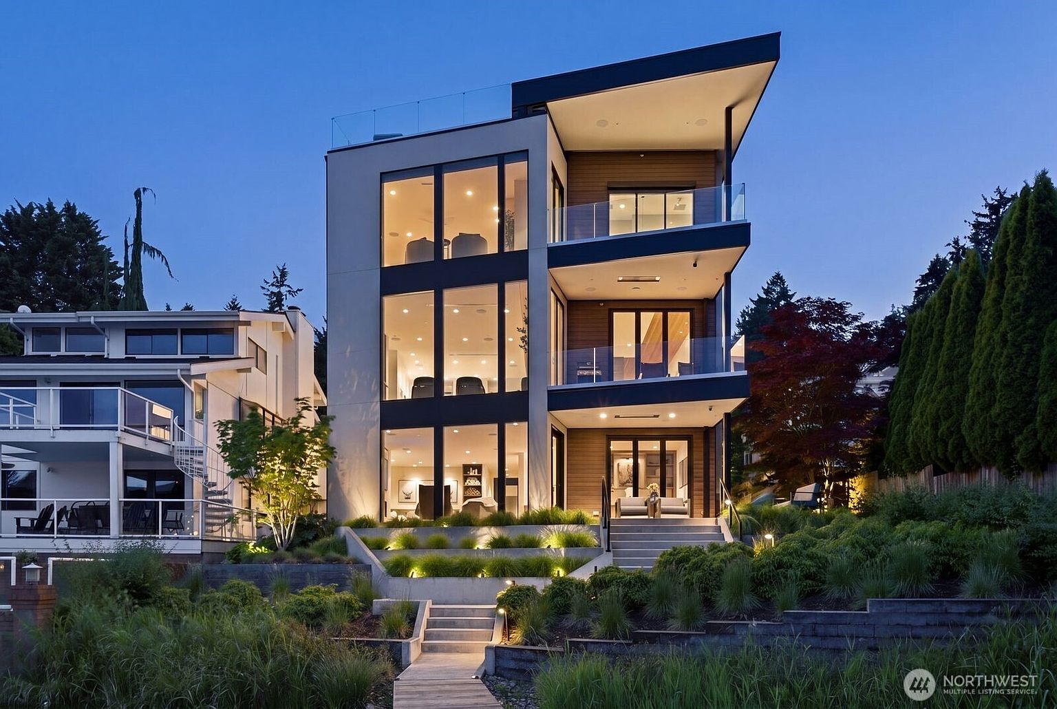 This striking modern residence features a multi-level design with expansive glass walls that offer a seamless connection between the interior and the outdoor environment. The home is characterized by its clean lines, tiered balconies with glass railings, and a sophisticated mix of light-toned siding and warm wood accents. Set against a twilight sky, the property is beautifully illuminated by integrated landscape lighting that highlights the terraced garden and stone pathways leading to the main entrance.