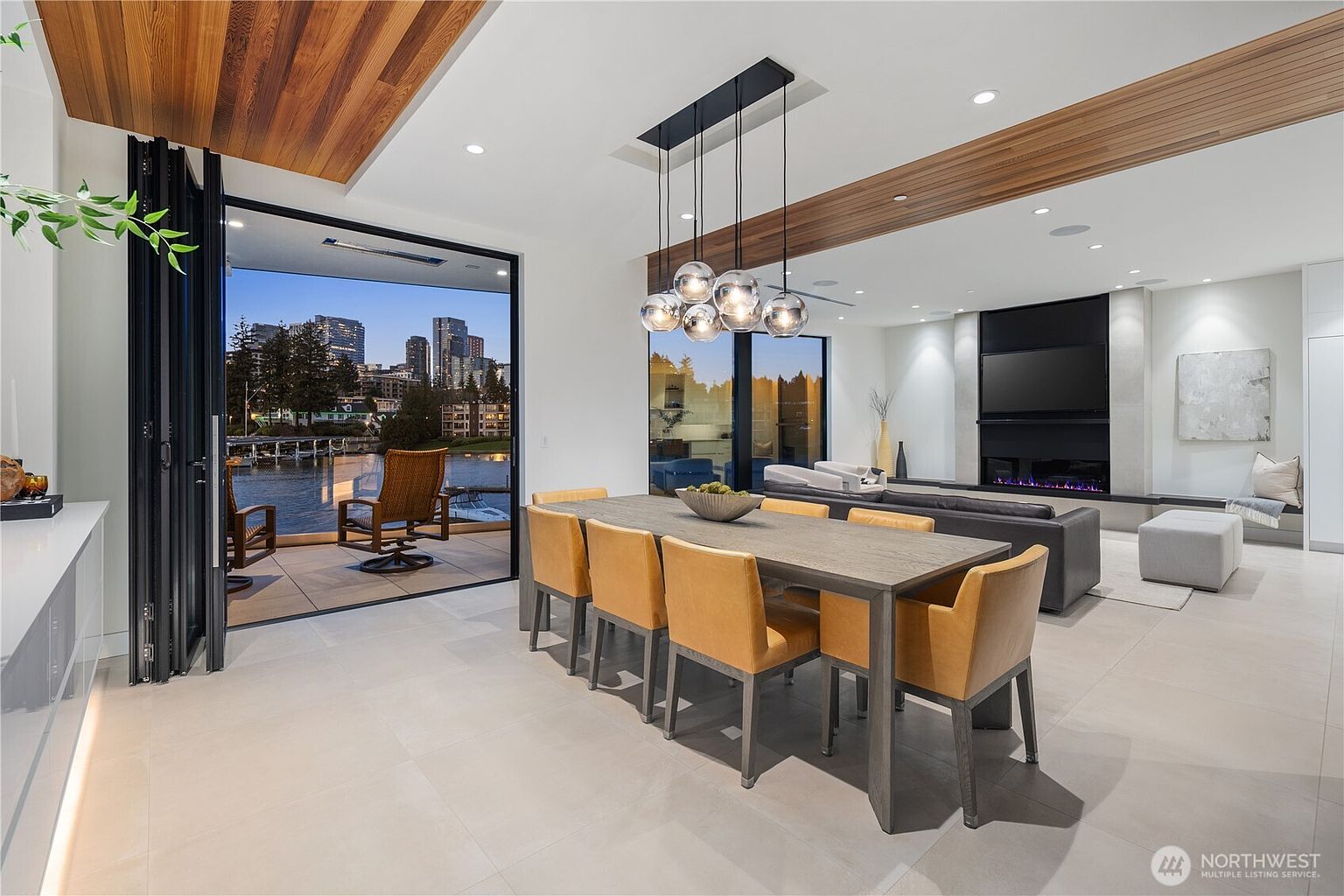 This open-concept dining area features a large, dark-toned wooden table surrounded by six tan leather chairs, illuminated by a modern cluster of glass globe pendant lights. The space flows seamlessly into a living area with a large fireplace and a wall-mounted television, while a wide set of folding glass doors opens to a covered patio overlooking a waterfront city view. The interior design is sleek and contemporary, characterized by neutral tile flooring, warm wood ceiling accents, and an expansive, airy atmosphere.