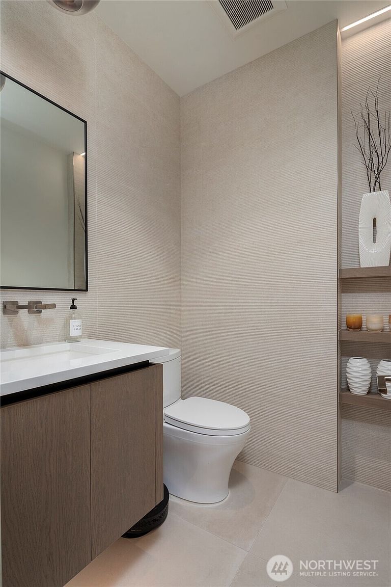 This modern powder room features textured, linear-patterned wall tiles that create a sophisticated, minimalist aesthetic. The space includes a floating vanity with a light wood finish, a sleek rectangular sink, and a wall-mounted faucet, complemented by a simple framed mirror. To the right, recessed shelving displays decorative vases and candles, adding a touch of warmth to the neutral, contemporary design.