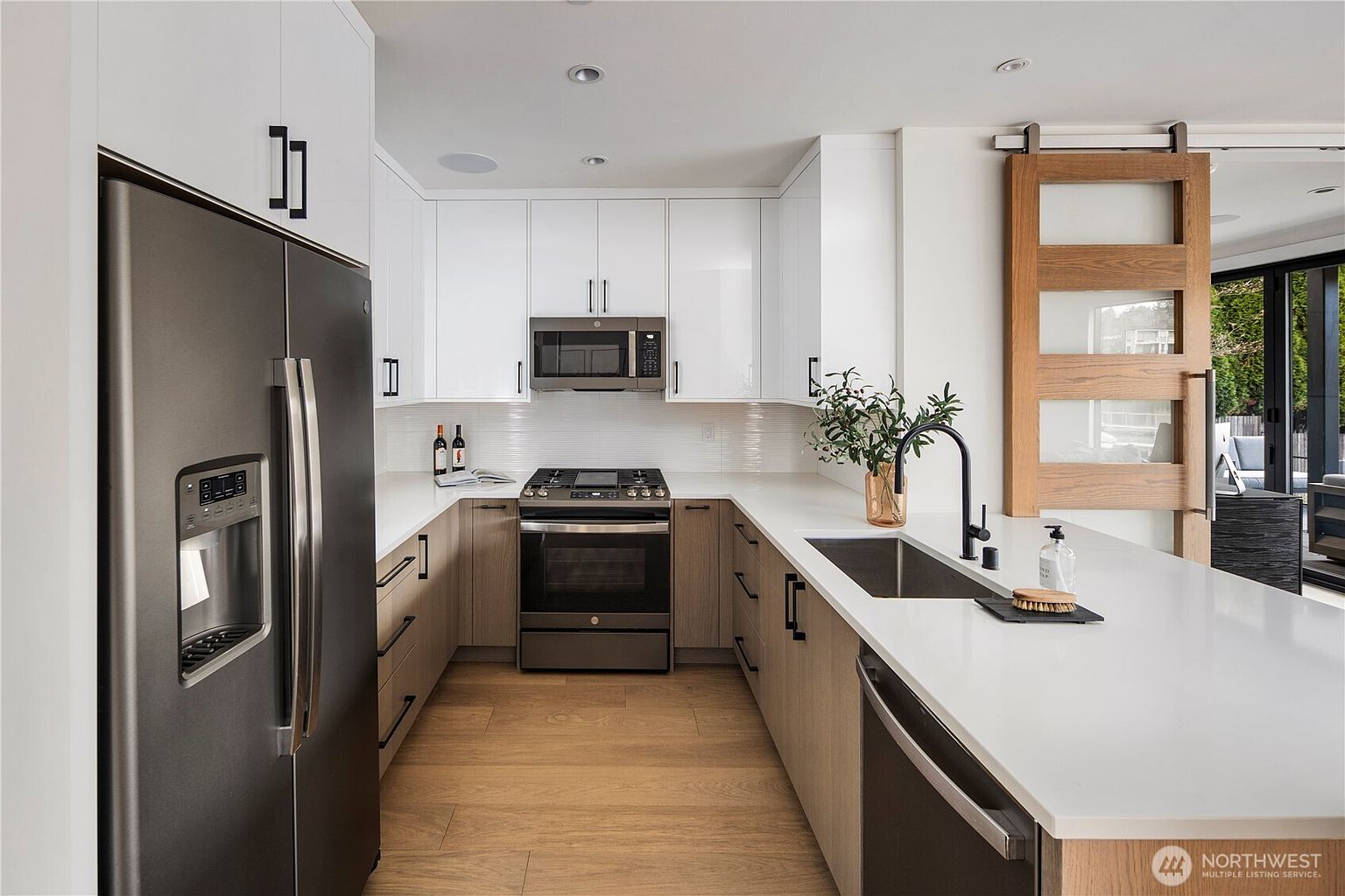 This modern, U-shaped kitchen features a clean aesthetic with white upper cabinets and light wood-toned lower cabinetry. The space is equipped with dark stainless steel appliances, a white countertop, and a sleek undermount sink with a matte black faucet. A sliding barn-style door with glass panels adds a rustic touch, while the perspective captures the functional layout from the kitchen island looking toward the range.