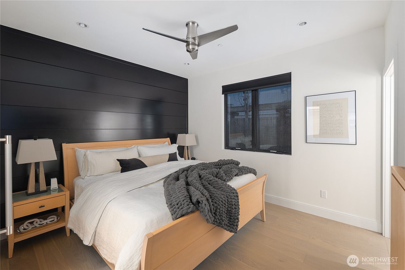 This modern bedroom features a striking black horizontal-plank accent wall behind a light wood bed frame. The room is brightened by neutral white walls, light wood flooring, and a large window with dark blinds. A contemporary ceiling fan and minimalist bedside lamps complete the clean, sophisticated aesthetic.