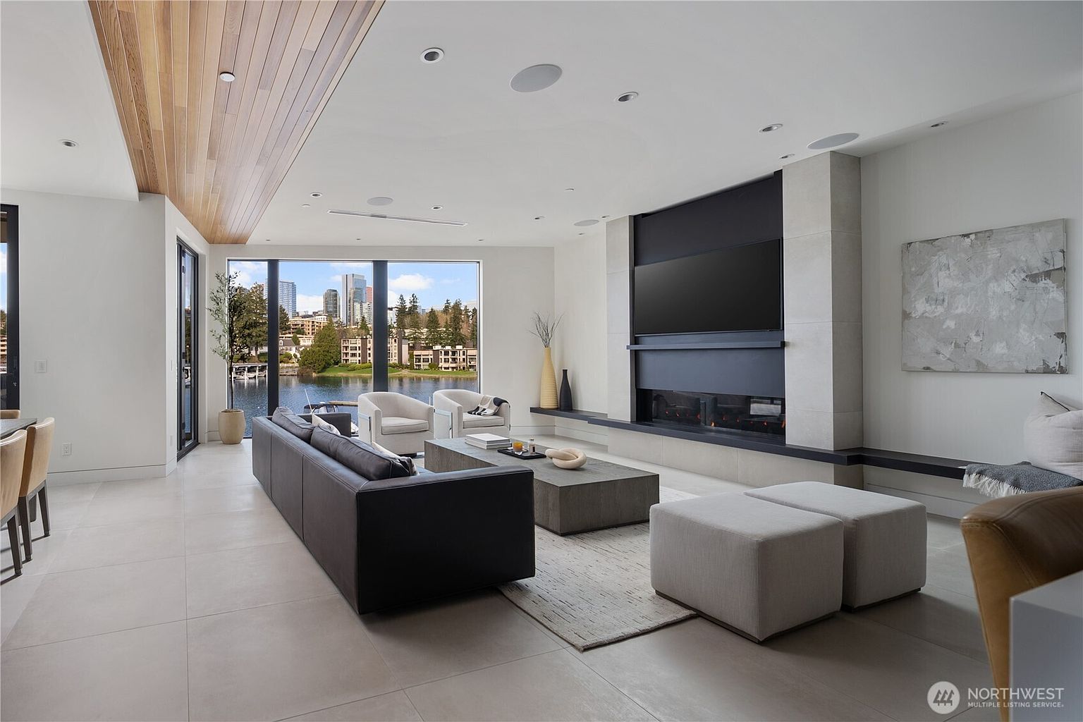 This contemporary living room features a spacious open-concept layout with large windows that frame a scenic waterfront view of a city skyline. The space is anchored by a dark leather sofa, a low-profile coffee table, and a prominent fireplace with a tall, light-toned stone surround and a black metal insert. A wood-paneled ceiling detail adds warmth to the neutral color palette, while the room is illuminated by recessed lighting and natural light.
