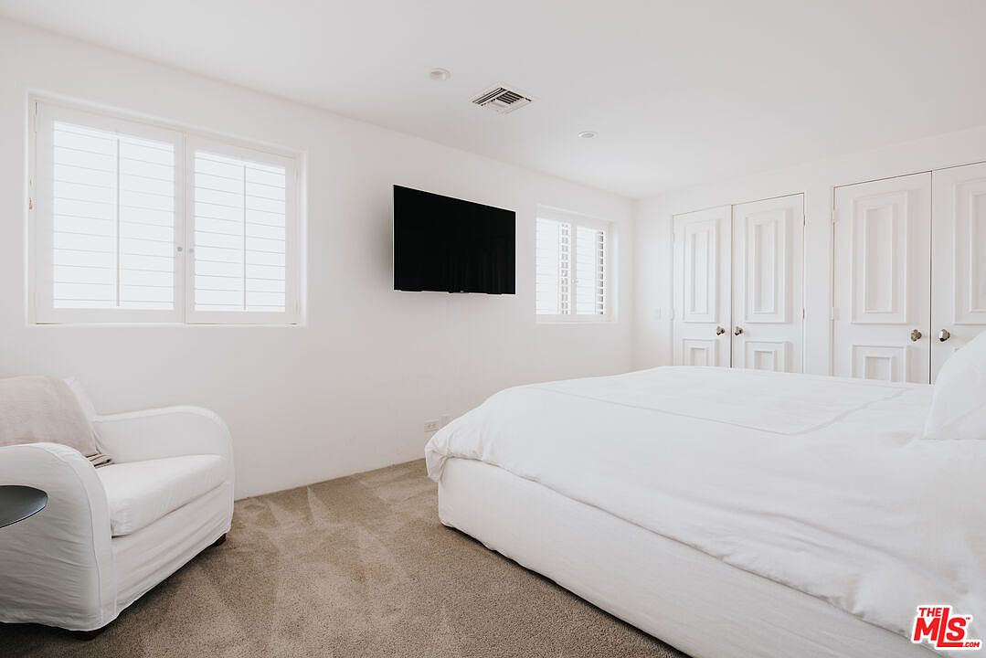 This bright and minimalist primary bedroom features a large, plush bed with white bedding as the central focus. The room is decorated in a clean, monochromatic white palette, complemented by soft beige carpeting and a comfortable white armchair in the corner. A wall-mounted television sits between two windows with plantation shutters, creating a serene and uncluttered atmosphere.