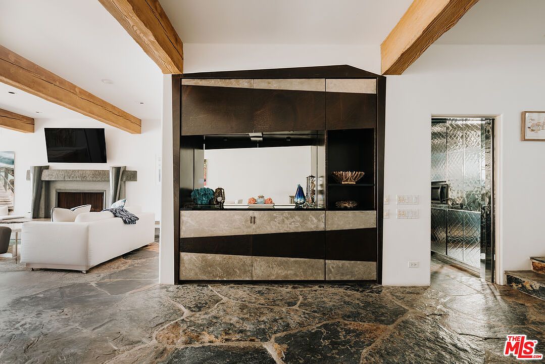 This interior shot captures a sophisticated living space featuring a striking custom built-in bar unit with geometric dark wood and stone paneling. The room is defined by rustic stone flooring and exposed wooden ceiling beams, creating a warm, textured aesthetic that flows into a cozy living area with a fireplace. The perspective is centered on the bar, showcasing a seamless blend of modern design and natural materials.