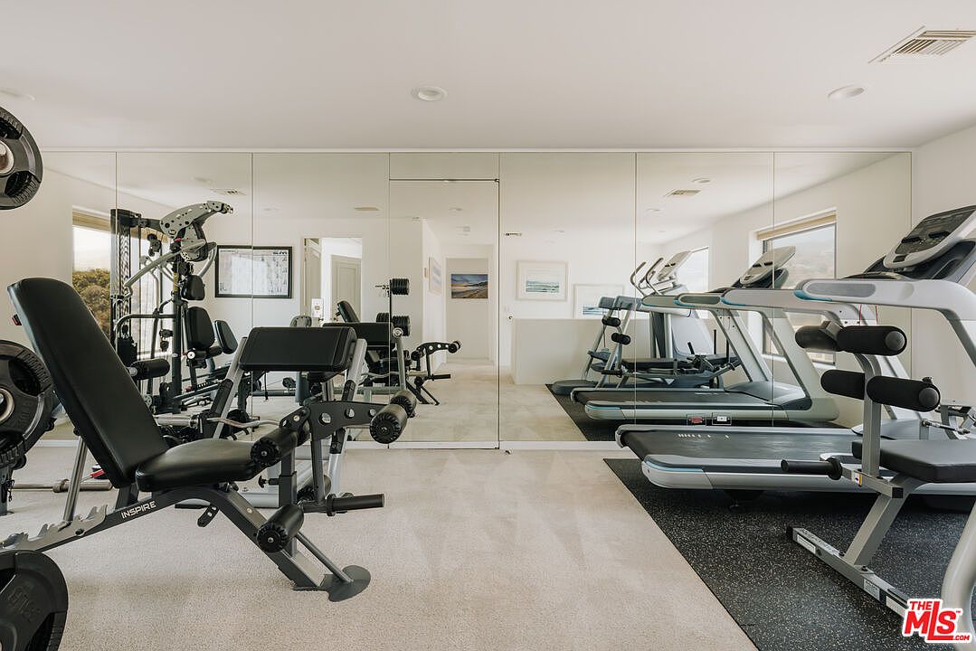 This well-equipped home gym features a variety of fitness equipment, including a multi-station weight machine and multiple treadmills positioned on a dedicated rubber mat. The room is enhanced by a full-length mirrored wall that creates a sense of spaciousness and reflects the bright, clean interior. The neutral color palette and functional layout make it an inviting space for a private workout.