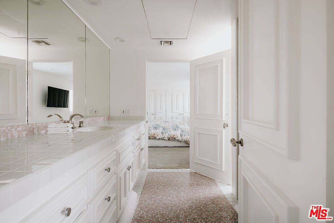 This bright, white-themed bathroom features a long vanity with a tiled countertop and a large wall-to-wall mirror, creating an expansive feel. The room leads directly into a bedroom, visible through the open doorway, showcasing a cohesive flow between spaces. The neutral color palette and clean lines provide a classic, well-maintained aesthetic suitable for a primary suite.