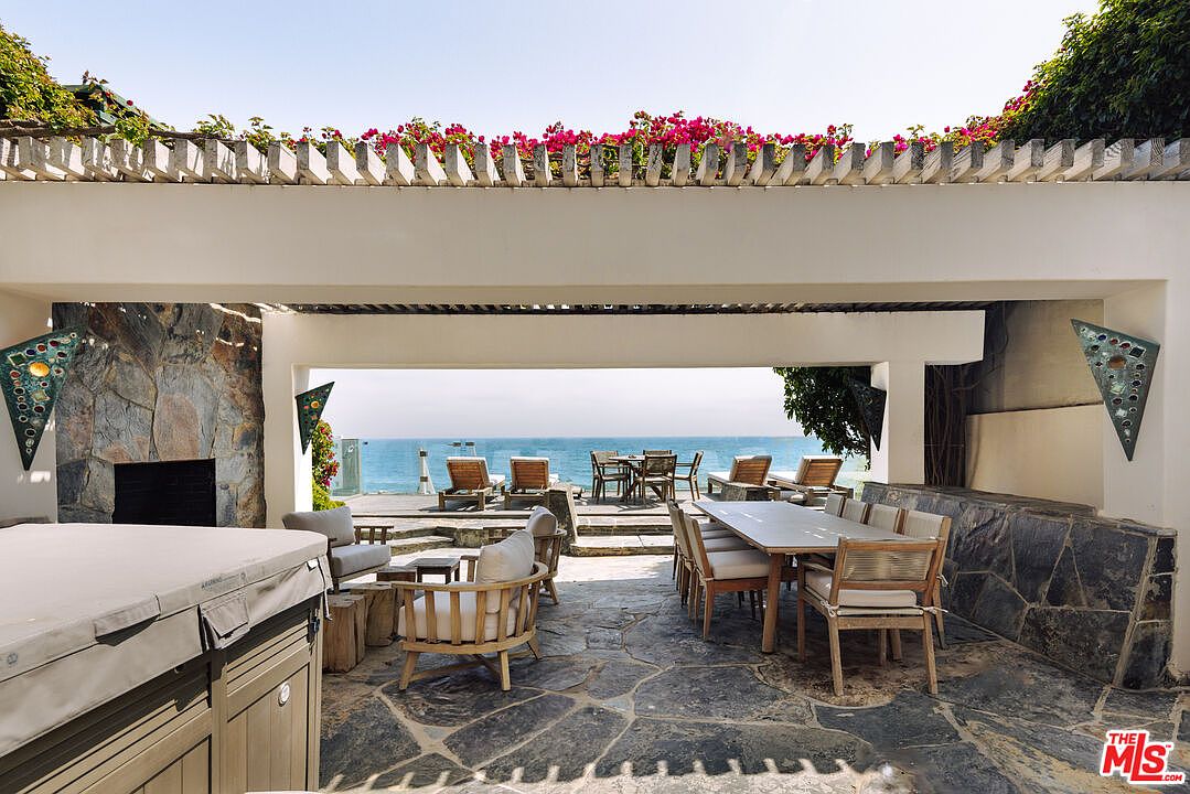 This expansive, covered outdoor patio offers a luxurious, open-air living space with direct, unobstructed views of the ocean. The area features a stone-paved floor, a built-in fireplace, a dining set, and comfortable lounge seating, all framed by a pergola adorned with vibrant bougainvillea. The seamless transition between the sheltered patio and the sun-drenched deck creates an ideal environment for high-end coastal entertaining.