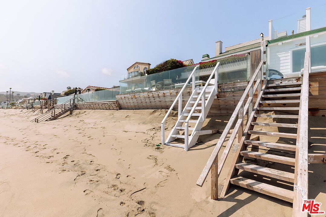 This image captures the rear exterior of a beachfront property, showcasing direct access to the sandy shore via multiple sets of wooden stairs. The home features a raised deck with glass railings, providing an unobstructed view of the ocean, while the architecture blends coastal charm with modern elements. The perspective is from the beach looking up, emphasizing the home's prime location and seamless transition between private living space and the natural environment.