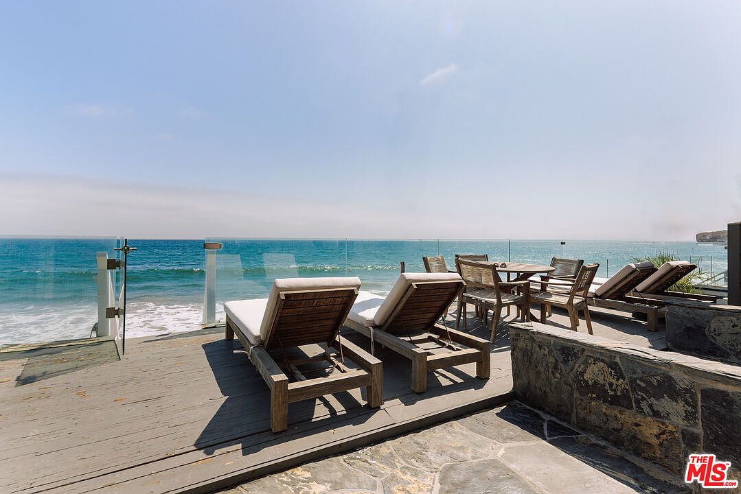 This stunning oceanfront deck features a weathered wooden floor and a glass railing that provides an unobstructed, panoramic view of the Pacific Ocean. The space is furnished with two comfortable lounge chairs in the foreground and a dining set further back, creating an ideal setting for relaxation or outdoor entertaining. The bright, sunny atmosphere and direct coastal access highlight the luxurious, serene lifestyle offered by this property.