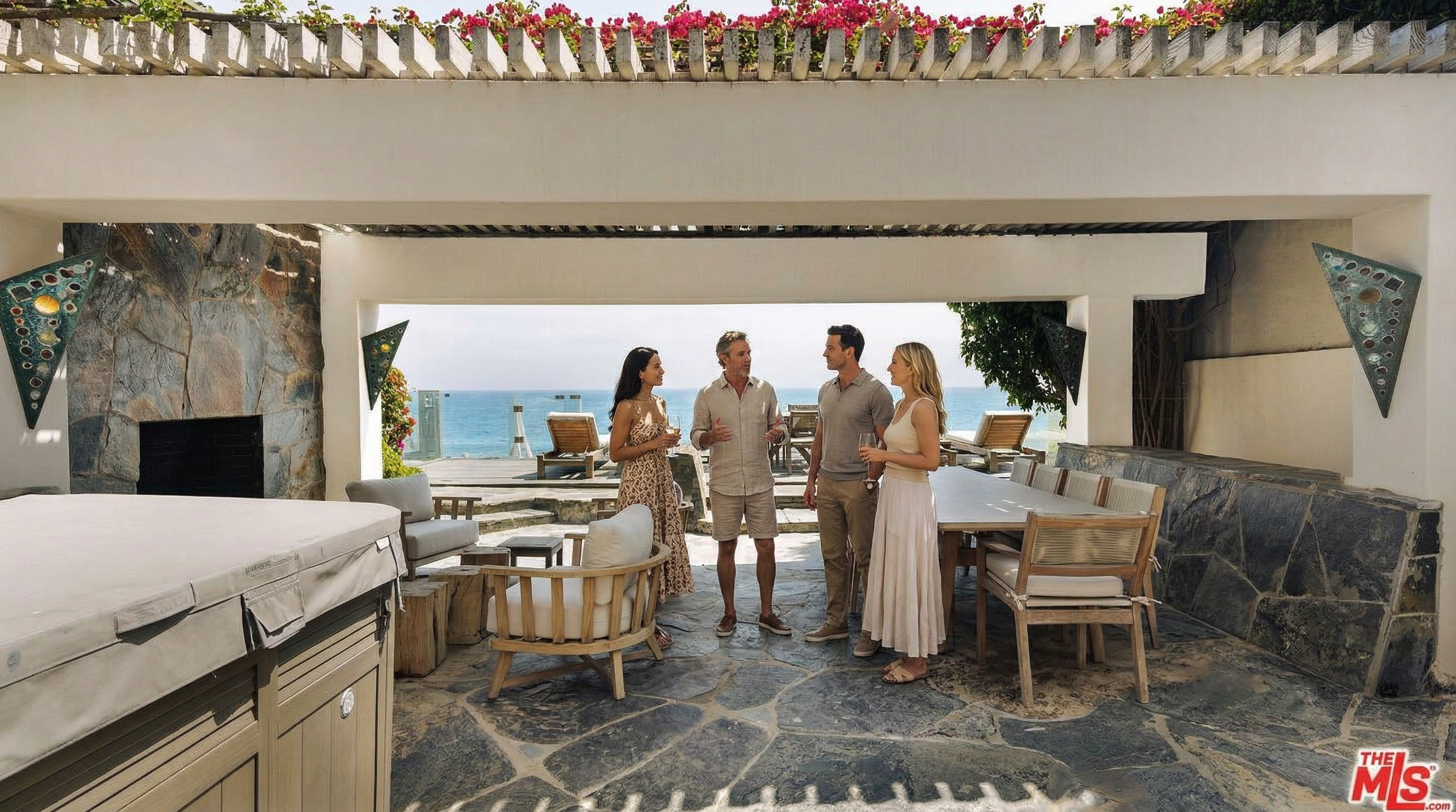 This expansive, covered outdoor patio offers a luxurious, open-air living space with direct, unobstructed views of the ocean. The area features a stone-paved floor, a built-in fireplace, a dining set, and comfortable lounge seating, all framed by a pergola adorned with vibrant bougainvillea. The seamless transition between the sheltered patio and the sun-drenched deck creates an ideal environment for high-end coastal entertaining.