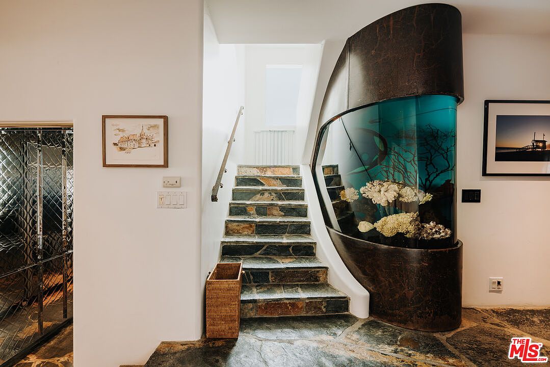 This unique interior space features a striking staircase constructed from natural stone, leading upward toward a bright, skylit landing. To the right, a large, custom-built curved aquarium serves as a dramatic architectural focal point, adding a vibrant, organic element to the home's design. The combination of the textured stone flooring, the sleek dark metal casing of the aquarium, and the clean white walls creates a sophisticated and artistic atmosphere.