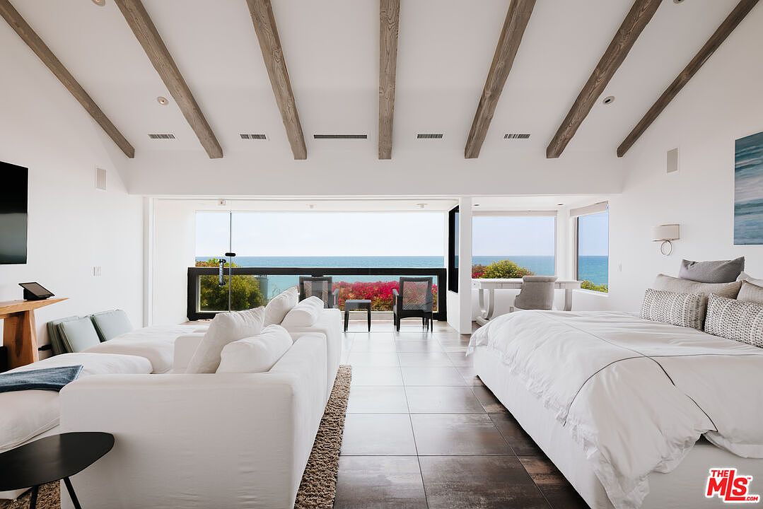 This expansive, light-filled primary bedroom features a seamless blend of modern luxury and coastal serenity, highlighted by vaulted ceilings with exposed wooden beams. The room is anchored by a plush white bed and a comfortable seating area, all oriented toward a massive floor-to-ceiling glass wall that offers an unobstructed, panoramic view of the ocean. The neutral color palette and tiled flooring create a sophisticated, airy atmosphere perfect for a high-end seaside retreat.