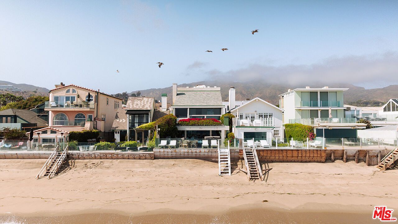 This eye-level shot captures a row of multi-story beachfront homes along a sandy coastline, showcasing their private decks and direct beach access. The architecture varies from traditional to contemporary, with each property featuring outdoor seating areas and staircases leading down to the sand. The backdrop features rolling hills under a bright, clear sky, emphasizing the desirable coastal lifestyle.