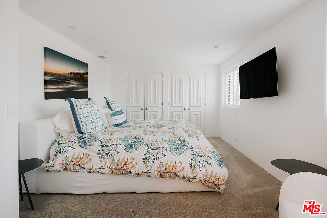 This bright and airy bedroom features a neutral color palette with a coastal-inspired coral and teal patterned duvet cover. The room is furnished with a plush bed, two small side tables, and a wall-mounted television, while double closet doors provide ample storage. The perspective is a straight-on shot that captures the clean, minimalist aesthetic and comfortable atmosphere of the space.