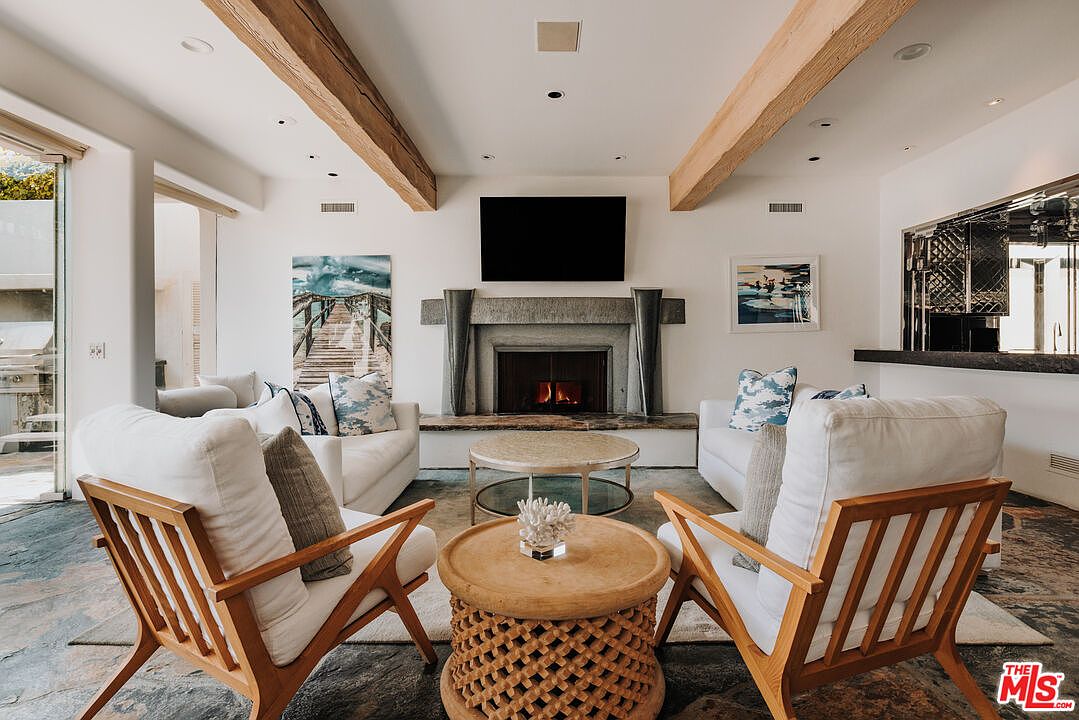 This elegant living room features a symmetrical layout centered around a striking stone fireplace with a mounted television. The space is defined by exposed wooden ceiling beams, white upholstered furniture, and a textured stone floor, creating a sophisticated yet relaxed coastal aesthetic. Large glass doors on the left invite natural light and provide a seamless transition to the outdoor patio area.