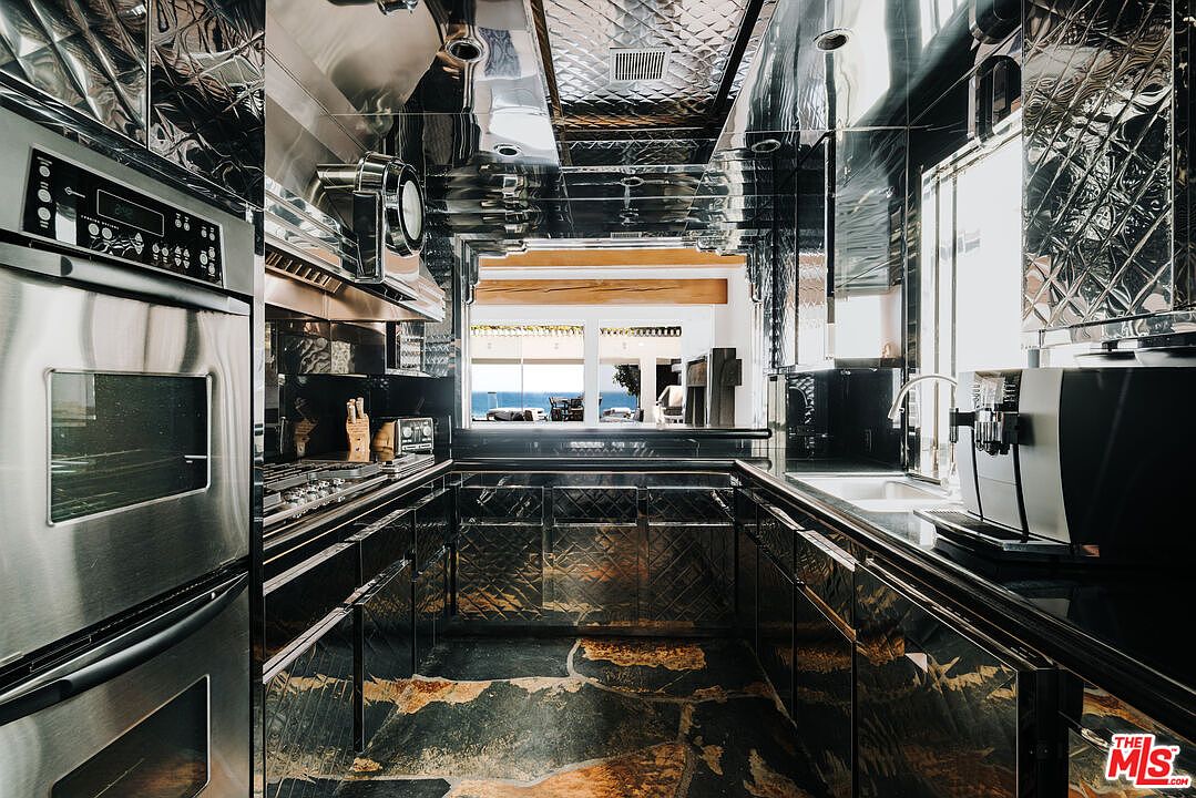This striking, high-contrast kitchen features a unique, reflective metallic aesthetic with diamond-patterned cabinetry and a polished, dark stone floor with gold veining. The space is equipped with professional-grade stainless steel appliances, including a built-in oven and a modern coffee machine, while a large window at the end of the galley layout provides a stunning, unobstructed view of the ocean. The overall impression is one of ultra-modern, luxurious, and dramatic design.