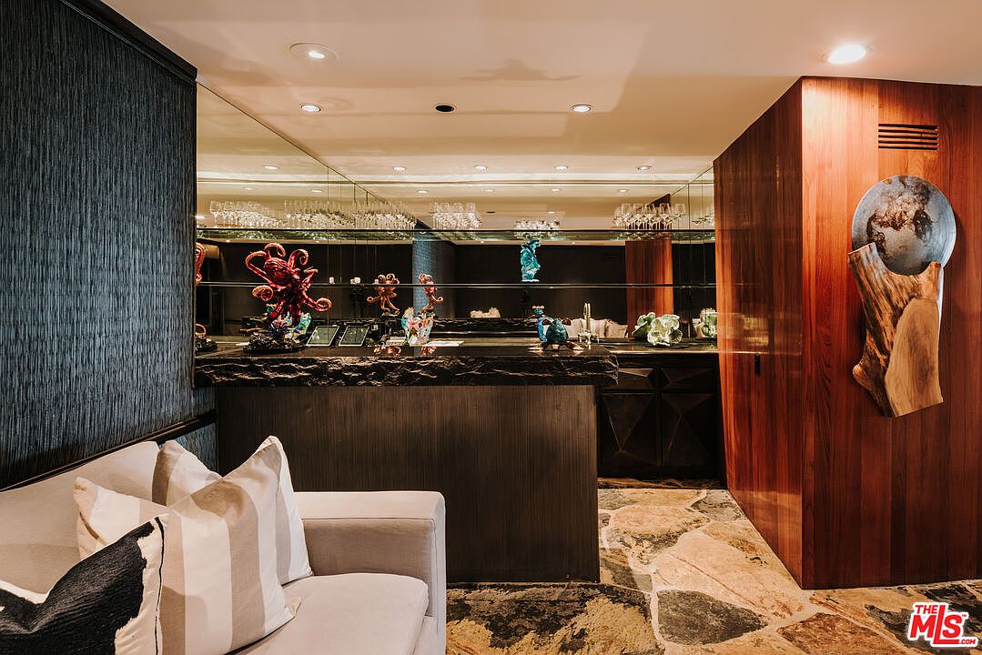 This sophisticated interior space features a custom wet bar with a rugged, dark stone countertop and a mirrored backsplash that reflects the room's elegant decor. A plush, light-colored sofa with patterned throw pillows sits in the foreground, while a striking wooden wall panel adorned with a circular art piece adds a warm, organic texture to the room. The combination of stone flooring, dark wood accents, and ambient recessed lighting creates a luxurious and moody atmosphere perfect for entertaining.