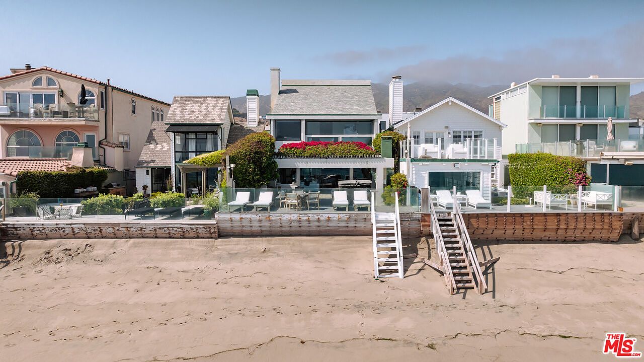 This beachside property features a row of multi-story homes directly overlooking the sandy shore, captured from a low-angle perspective on the beach. The central residence showcases a modern aesthetic with a prominent second-story deck adorned with vibrant red flowers, while wooden staircases provide direct access from the elevated patios down to the sand. The scene conveys a serene, upscale coastal lifestyle with clear views of the oceanfront architecture and the hazy mountains in the background.