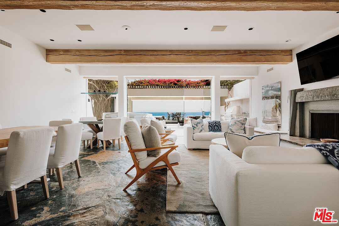 This expansive, open-concept living and dining area features a rustic aesthetic with exposed wooden ceiling beams and a textured stone floor. The space is anchored by a cozy fireplace and plush white seating, seamlessly transitioning to an outdoor patio with ocean views through large glass doors. The bright, airy atmosphere is enhanced by neutral tones and natural light, creating a sophisticated yet relaxed coastal living environment.