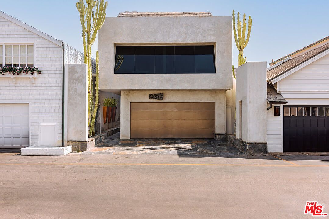 This striking modern residence features a minimalist, boxy architectural design with a smooth, light-colored stucco facade and a prominent, wide horizontal window. The ground level is dominated by a large, metallic-finished garage door, flanked by tall, sculptural cacti that add a unique, desert-modern aesthetic. The property is situated between two more traditional homes, creating a bold contrast in style, and the stone-paved driveway leads directly to the street.