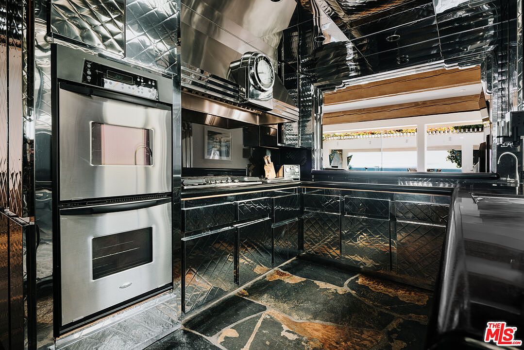 This unique, high-contrast kitchen features a striking Art Deco-inspired aesthetic with reflective, diamond-patterned metallic cabinetry and matching mirrored surfaces. The space is equipped with professional-grade stainless steel double ovens and a built-in range hood, set against a rugged, dark stone floor. A large pass-through window provides a view into an adjacent living area, creating an open and dramatic atmosphere.