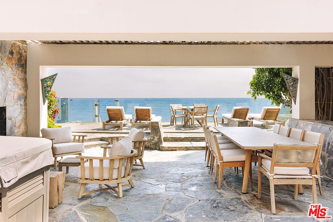 This expansive, open-air patio offers a seamless transition between indoor living and the stunning oceanfront view. The space is furnished with a large dining set and comfortable lounge seating, all set against a backdrop of natural stone flooring and a panoramic sea vista. The architectural design emphasizes an airy, Mediterranean-inspired aesthetic perfect for outdoor entertaining.