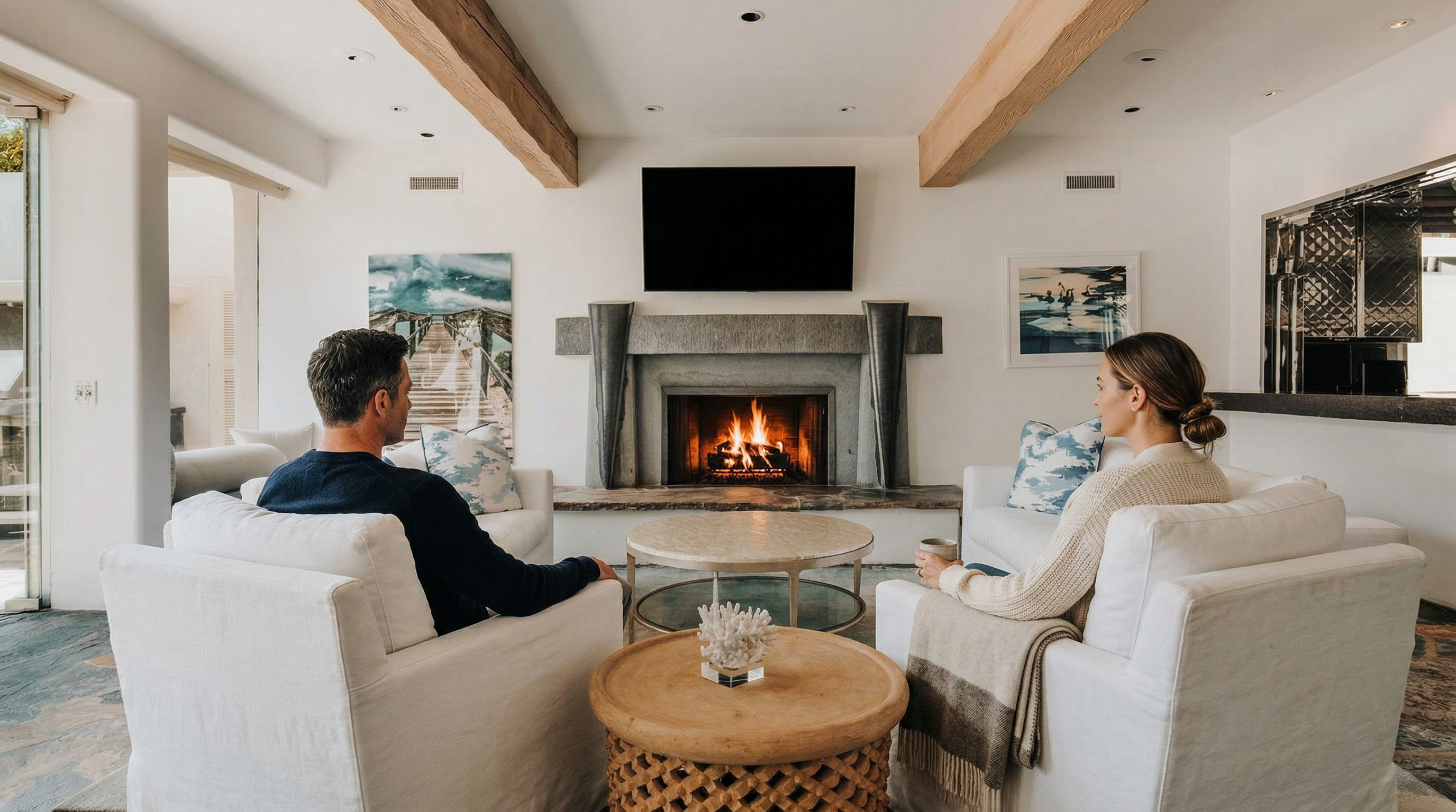 This elegant living room features a symmetrical layout centered around a striking stone fireplace with a mounted television. The space is defined by exposed wooden ceiling beams, white upholstered furniture, and a textured stone floor, creating a sophisticated yet relaxed coastal aesthetic. Large glass doors on the left invite natural light and provide a seamless transition to the outdoor patio area.