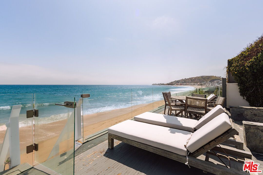 This stunning oceanfront deck offers a seamless transition between indoor living and the Pacific coastline. The space is furnished with two comfortable lounge chairs and a dining set, all positioned to maximize the panoramic beach and ocean views. A sleek glass railing ensures an unobstructed perspective, creating a serene and luxurious atmosphere perfect for coastal relaxation.
