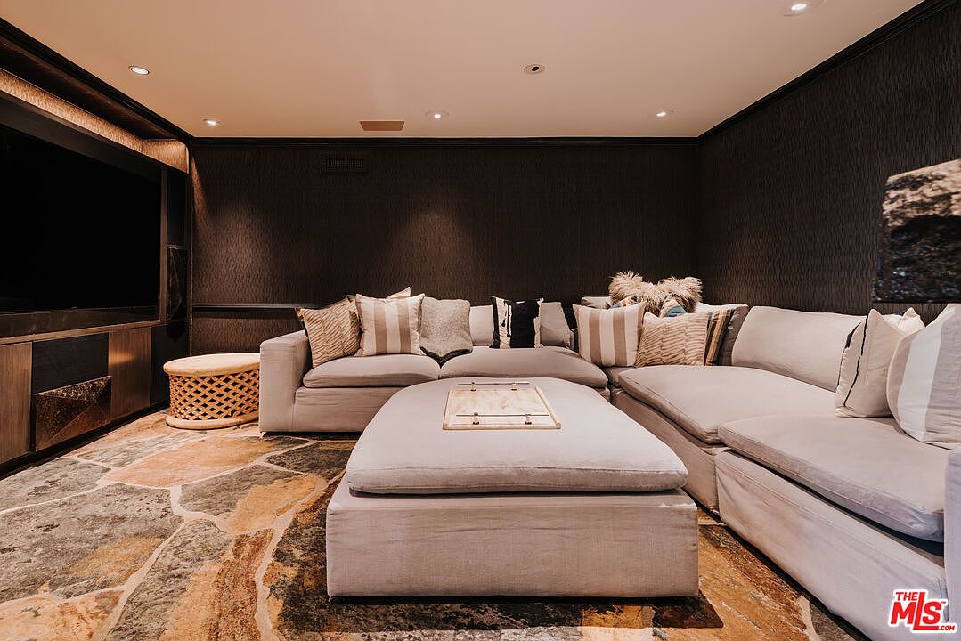 This cozy, dark-toned media room features a large, plush sectional sofa with a matching oversized ottoman, perfect for lounging. The space is anchored by a unique stone-patterned area rug and a built-in entertainment center, creating a sophisticated and intimate atmosphere. The low-angle perspective emphasizes the comfort and scale of the seating area, highlighting it as an ideal space for relaxation or home cinema.