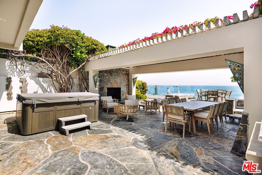 This expansive, stone-paved outdoor patio offers a luxurious coastal living experience, featuring a private hot tub, a cozy fireplace seating area, and a formal dining space. The open-air design is framed by a pergola adorned with vibrant bougainvillea, providing an unobstructed, panoramic view of the ocean. The combination of natural stone, elegant outdoor furniture, and the serene seaside backdrop creates an inviting and sophisticated atmosphere for relaxation or entertaining.