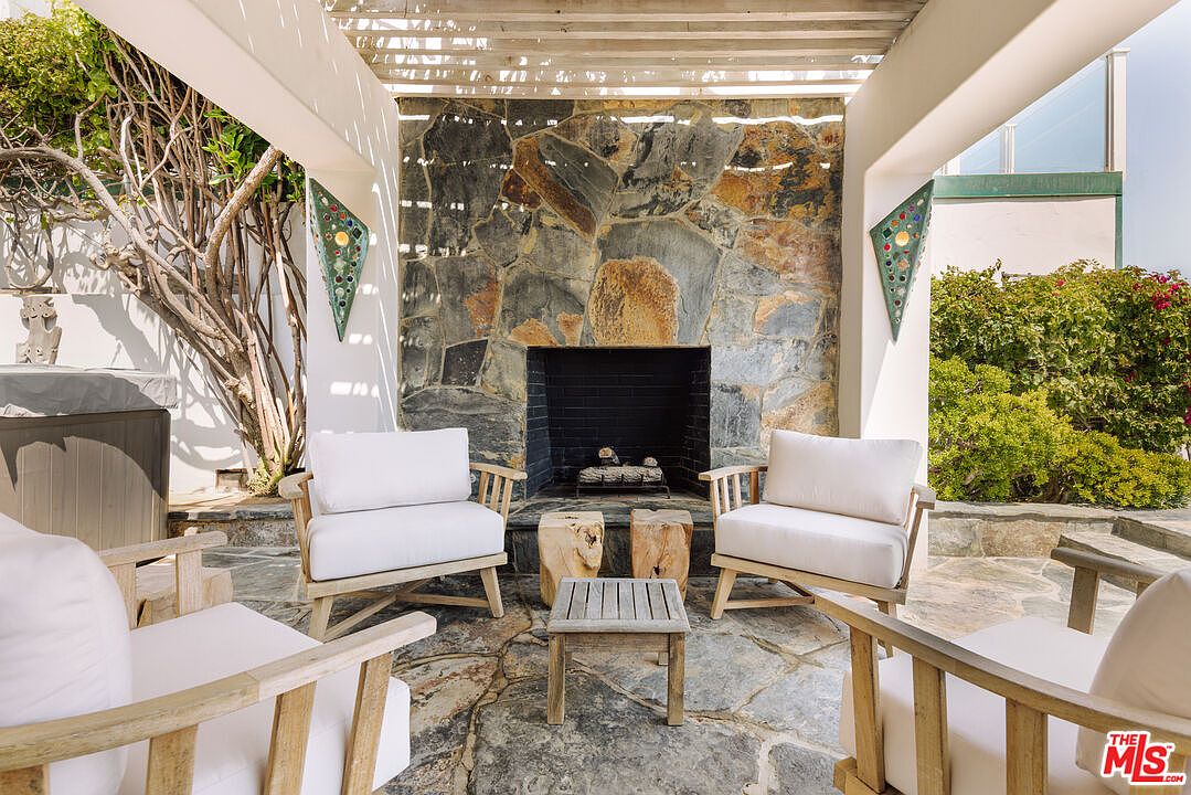This inviting outdoor patio features a rustic stone fireplace as its centerpiece, framed by a wooden pergola overhead. The space is furnished with comfortable white-cushioned armchairs and natural wood side tables, creating a cozy and relaxed seating area on a flagstone floor. The setting is enhanced by lush greenery and decorative wall sconces, offering a serene and sophisticated atmosphere perfect for outdoor entertaining.