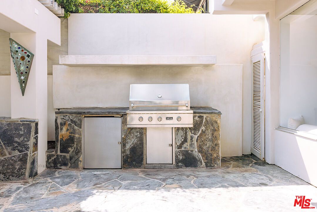 This outdoor kitchen area features a built-in stainless steel grill and a matching under-counter refrigerator set into a rustic stone-clad base. The space is framed by a clean, white stucco wall with a floating shelf above the grill, creating a modern yet textured aesthetic. The stone flooring and natural light contribute to an inviting, high-end outdoor entertaining atmosphere.