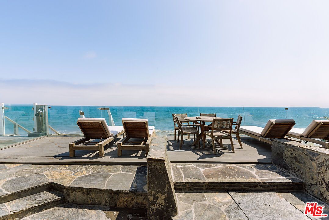 This expansive oceanfront deck offers a serene and luxurious outdoor living space, featuring a combination of stone flooring and wooden decking. The area is furnished with multiple lounge chairs and a central dining set, all positioned to maximize the panoramic views of the sea. A sleek glass railing ensures an unobstructed connection to the horizon, creating an inviting atmosphere for relaxation or entertaining.