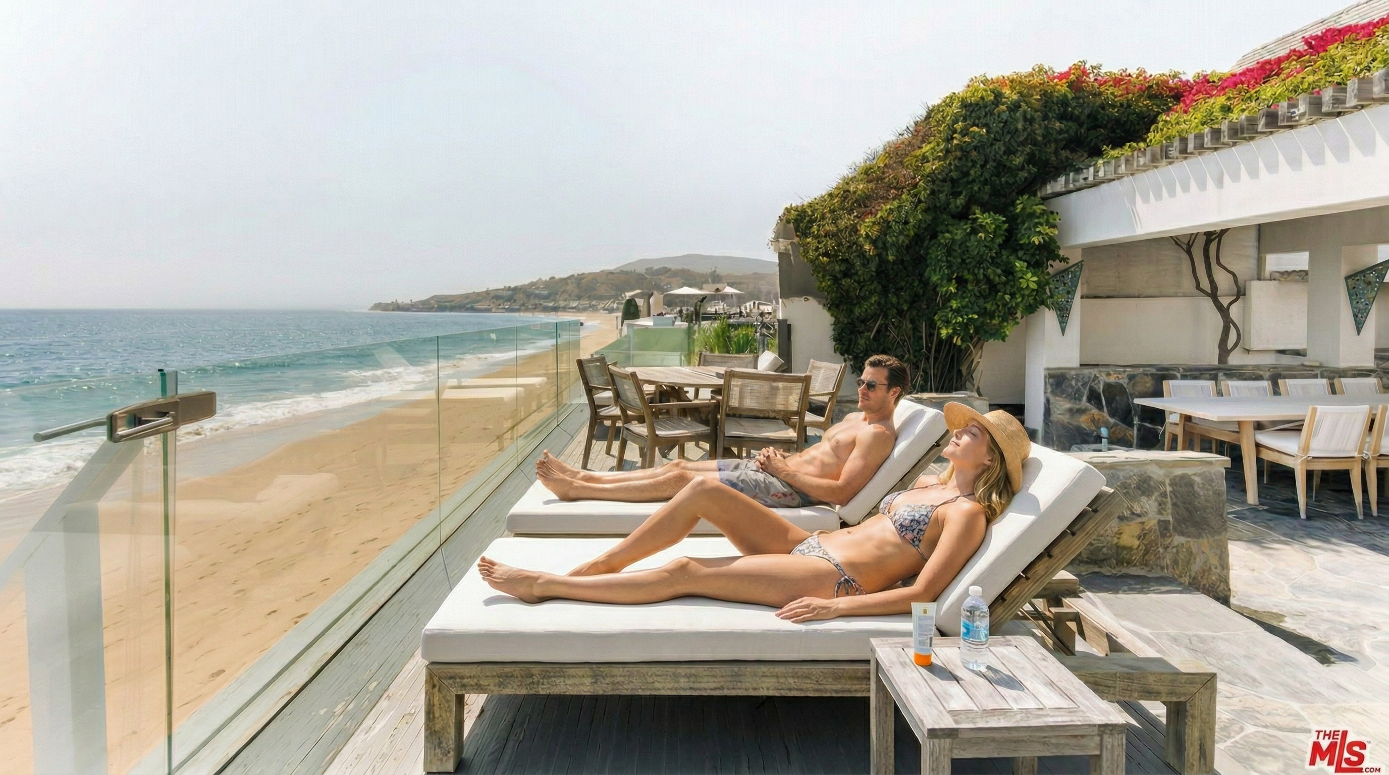 This stunning oceanfront deck offers a luxurious outdoor living space with direct, unobstructed views of the beach and coastline. The area is furnished with comfortable wooden lounge chairs and a dining set, all set against a backdrop of lush greenery and a modern glass railing. The perspective captures the seamless transition between the private terrace and the expansive sandy beach, evoking a serene and high-end coastal lifestyle.