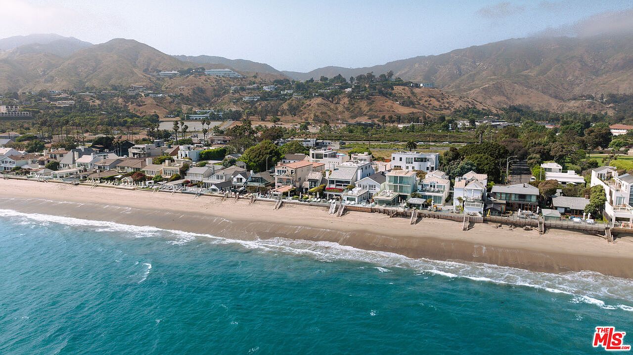 This high-angle aerial shot captures a stunning stretch of beachfront property along a coastal highway, showcasing a row of multi-story homes directly overlooking the ocean. The scene features a sandy beach, rolling waves, and a backdrop of lush, mountainous terrain under a clear sky. The perspective emphasizes the prime location and the seamless integration of luxury residential living with the natural beauty of the coastline.