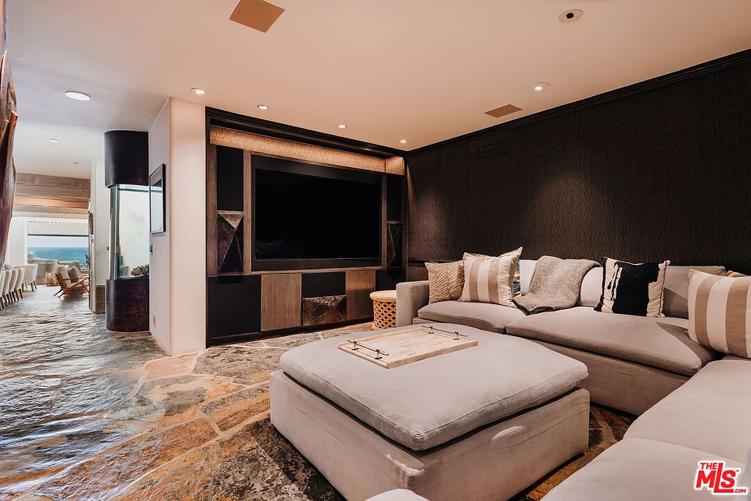 This sophisticated living room features a large, plush sectional sofa with a matching ottoman, centered around a sleek, dark-toned media wall with built-in cabinetry. The room is anchored by unique, textured stone flooring that extends into an adjacent hallway, creating a seamless flow toward a bright, ocean-view opening. The overall aesthetic is modern and moody, utilizing dark wall textures and warm recessed lighting to create an intimate, high-end atmosphere.