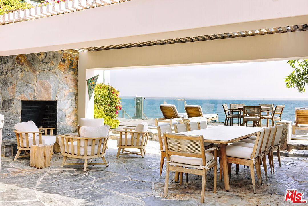 This expansive, open-air patio offers a luxurious outdoor living space with a stone fireplace and a dining area overlooking the ocean. The space features natural wood furniture with white cushions, a rustic stone floor, and a covered ceiling with wooden slats, creating a seamless blend of indoor comfort and coastal scenery. The panoramic view of the sea provides a serene and high-end atmosphere perfect for entertaining.