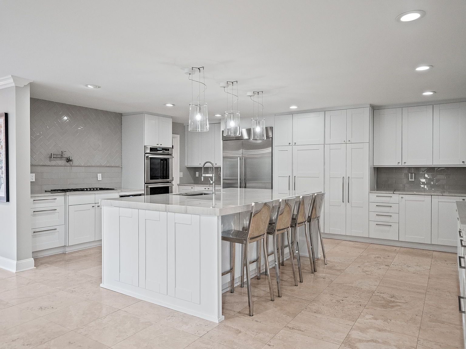This is a bright and spacious kitchen featuring white cabinetry, stainless steel appliances, and a large center island with a marble countertop. The kitchen is well-lit with recessed lighting and pendant lights above the island, creating a clean and modern aesthetic. The floor is tiled with a light beige stone, adding warmth to the space.