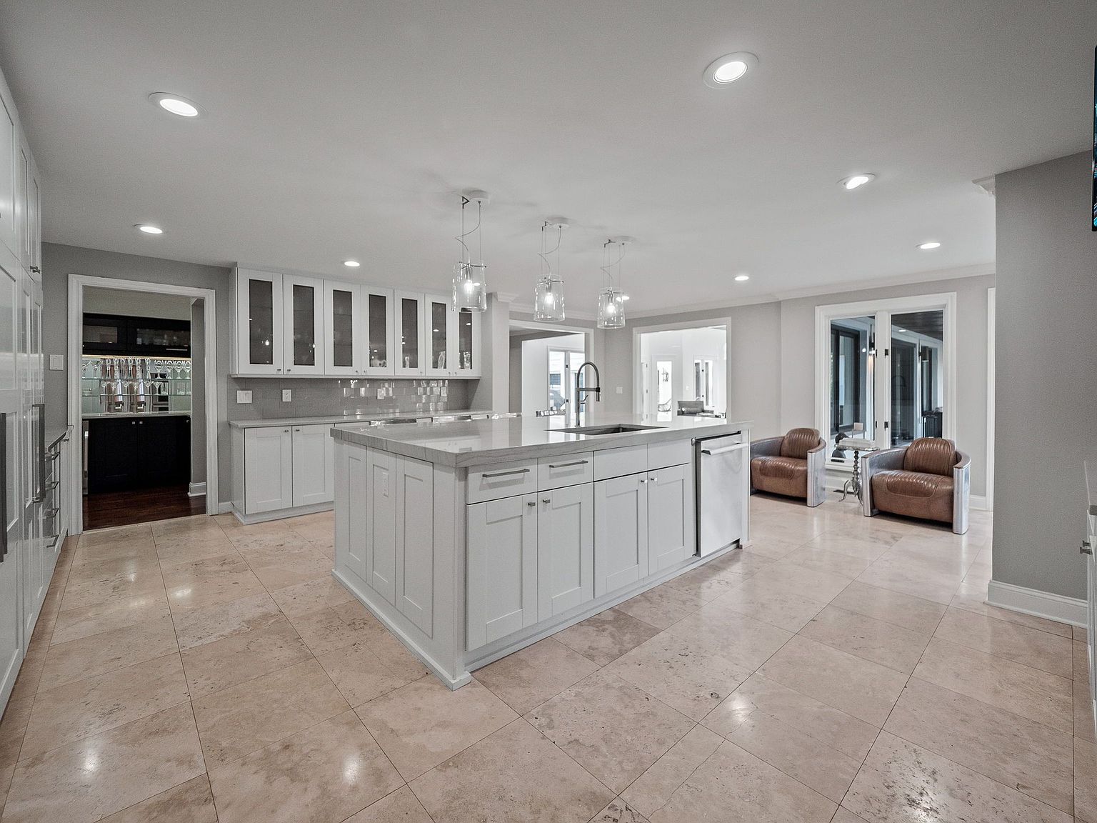This is a bright and spacious kitchen featuring a large central island with a sink and white cabinetry. The kitchen includes stainless steel appliances and pendant lighting above the island. The flooring is a light-colored tile, and there is a bar area visible through a doorway, as well as a seating area with armchairs near a set of glass doors.